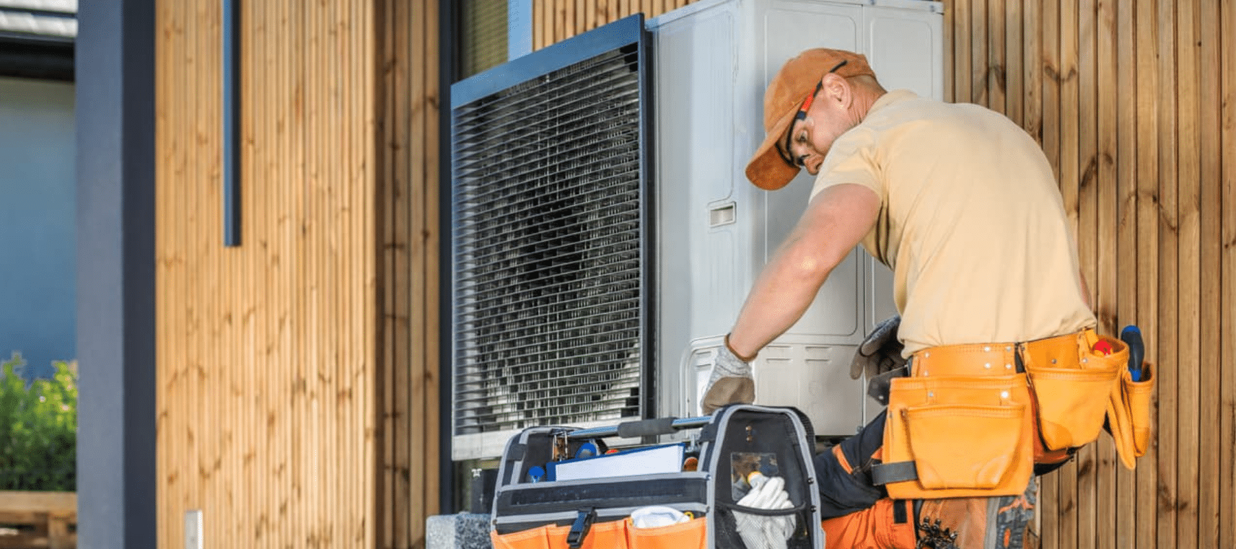 A maintenance worker inspecting and servicing an outdoor HVAC system near a modern wooden building