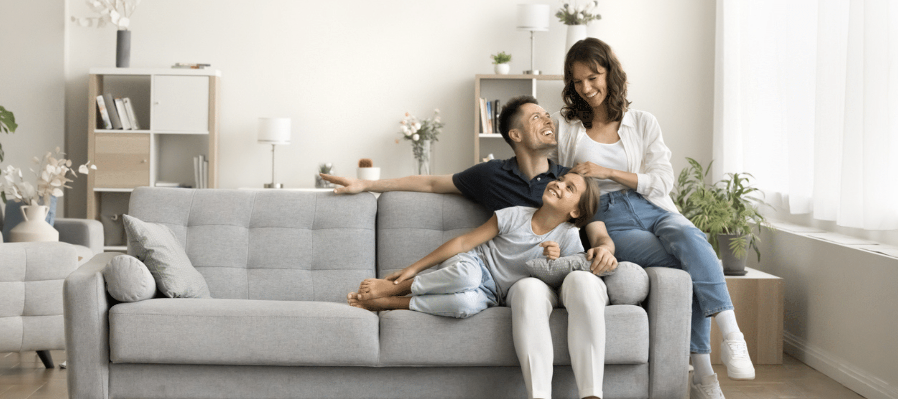 A happy family of three—father, mother, and daughter—relaxing together on a gray couch in a bright, modern living room